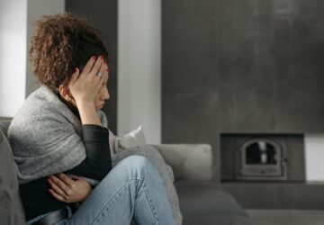 Kobieta z ręką na głowie, siedząca na kanapie, wyglądająca na zmęczoną i zmartwioną