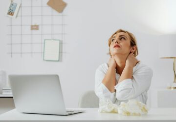 Kobieta siedząca przy biurku, z rękami na szyi, patrząca w górę, z użytymi chusteczkami obok laptopa