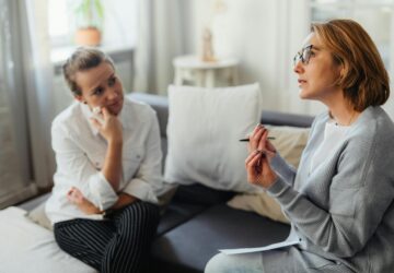 Dwie kobiety rozmawiające na kanapie, jedna z nich mówi, trzymając długopis, a druga słucha