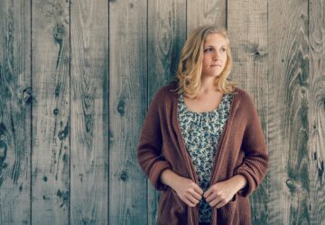 Kobieta stojąca przed drewnianą ścianą, z rękami w kieszeniach swetra