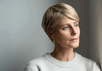 Kobieta o krótkich blond włosach patrzy zamyślona przez okno.