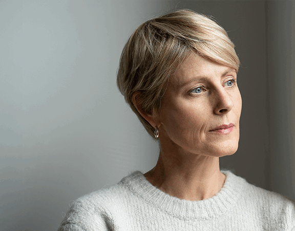 Kobieta o krótkich blond włosach patrzy zamyślona przez okno.