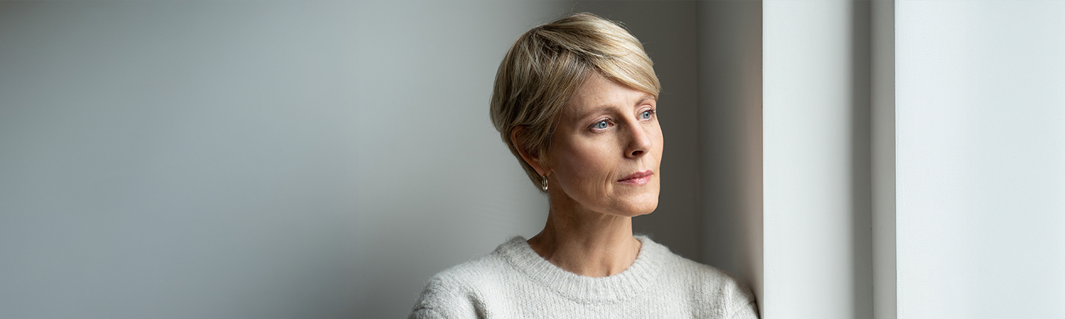 Kobieta o krótkich blond włosach patrzy zamyślona przez okno.