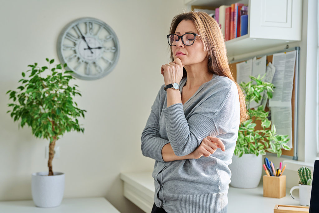 Kobieta w okularach stoi zamyÅ›lona z rÄ™kÄ… przy brodzie.